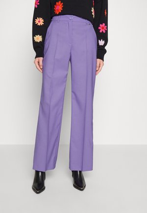 Frau trägt hoch taillierte, gerade geschnittene Lavendelhose, schwarzen floralen Pullover und schwarze spitze Stiefel, steht vor einfachem Hintergrund.