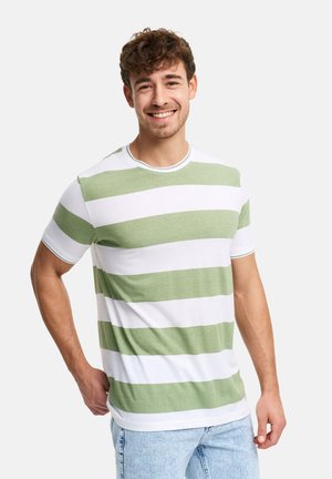 Joven sonriente, vistiendo una camiseta de rayas horizontales verdes y blancas y jeans azul claro, de pie contra un fondo liso.