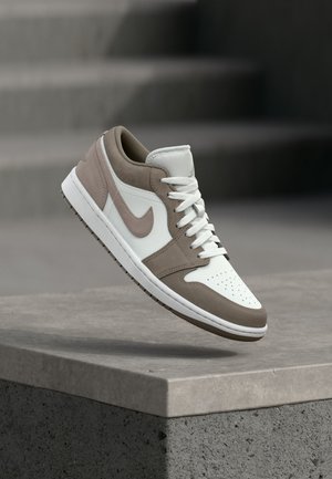 White and taupe low-top sneaker with laces, midsole, and Nike swoosh hovering above a concrete ledge with stairs in background.