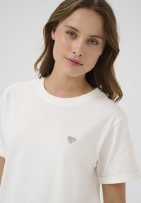 Cream cotton t-shirt featuring a small, sparkling heart embellishment on the left chest, with rolled sleeve cuffs and a classic crew neckline.