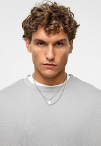 Grijze sweatshirt met een ronde hals, gemaakt van een gestructureerde stof. Twee zilveren kettingen met een rechthoekige hanger lagen over een wit T-shirt.