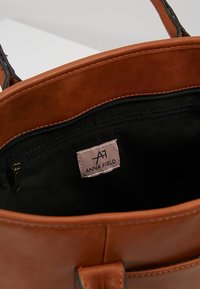 Indvendig brun taske i kunstlæder med sort foer, der har en lynlåslomme og en "ANNA FIELD"-mærkat. Rundet design med synlige syninger.