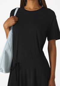 Schwarzes T-Shirt-Kleid mit kurzen Ärmeln, das eine geraffte Taille mit Schnürdetail aufweist. Eine hellblaue Schultertasche wird über die Schulter gehalten.