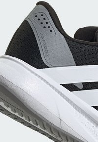 Scarpa sportiva nera con tomaia testurizzata, dettaglio grigio sul tallone e suola in gomma bianca con un motivo del battistrada ben definito. Struttura leggera evidente.