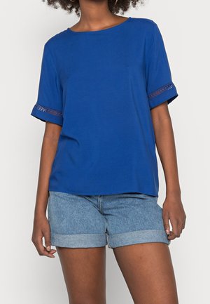 Kvinde iført en blå top med korte ærmer med blonder på ærmerne og lyse, op rullede denimshorts mod en ensfarvet baggrund.