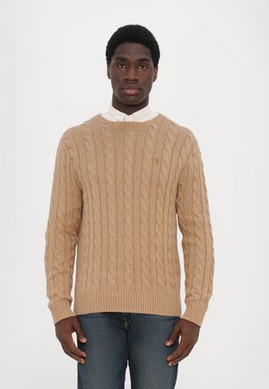 Beige kabelstrikket sweater med ribbede halsudskæring og manchetter. Har et struktureret, snoet mønster, lagdelt over en hvid kravebluse.
