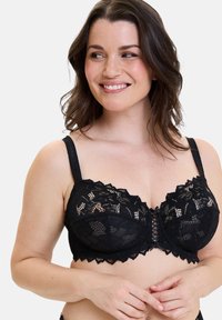 Mujer sonriente con cabello castaño hasta los hombros que lleva un sujetador negro de encaje con motivos florales y tirantes ajustables.