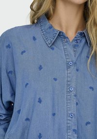 Camicia di denim azzurro con colletto, caratterizzata da ricami floreali blu sparsi e bottoni chiari lungo la parte anteriore. Tessuto morbido.