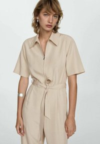 Salopette beige a maniche corte con zip frontale e vita con lacci. Presenta un colletto e una texture liscia. Design minimalista senza motivi.