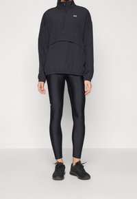 Czarna sportowa kurtka na zamek, o luźnym kroju, z warstwową konstrukcją i marszczonym dołem, zestawiona z błyszczącymi czarnymi legginsami i czarnymi trampkami.