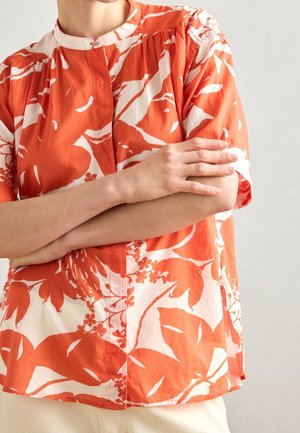 Osoba ubrana w pomarańczowo-białą krótkorękawową bluzkę w kwiaty z kołnierzykiem typu mandarynka, z założonymi na piersi skrzyżowanymi ramionami.