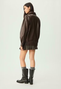 Giacca oversize in finta pelle marrone con colletto, polsini a coste, abbinata a una gonna plissettata e stivali neri al ginocchio.
