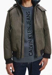 Kurtka typu bomber w oliwkowej zieleni o teksturowanym wykończeniu, z suwakiem z przodu i ściągaczami przy rękawach. Noszona na ciemnoszarym swetrze z logo. Na dole casualowe dżinsy.