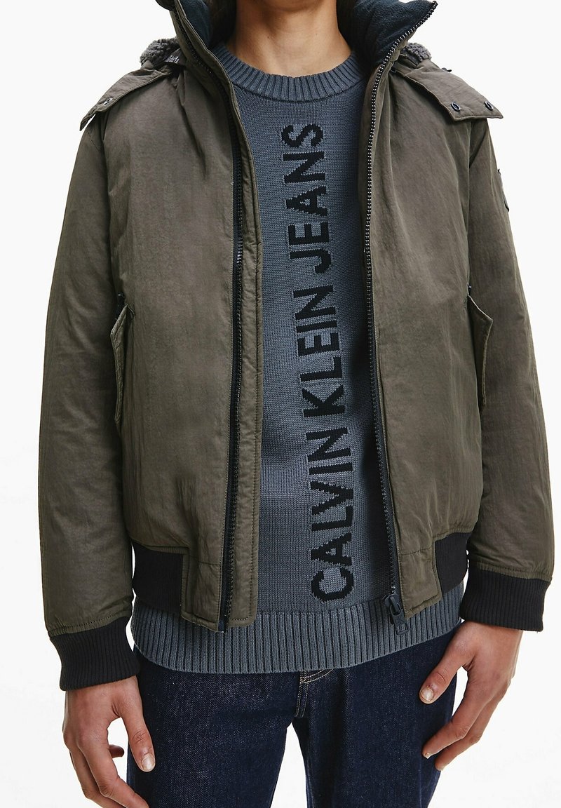 Kurtka typu bomber w oliwkowej zieleni o teksturowanym wykończeniu, z suwakiem z przodu i ściągaczami przy rękawach. Noszona na ciemnoszarym swetrze z logo. Na dole casualowe dżinsy.