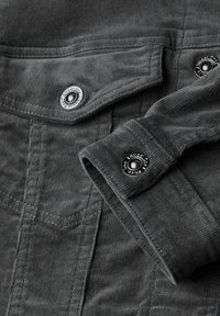 Šedá kordurová látka s viditelnou texturou jemných žeber, opatřená zesílenými švy a kovovými patentními knoflíky na klopě a manžetách.