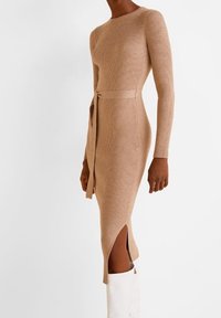 Robe en tricot côtelé beige avec des manches longues, une silhouette ajustée, une ceinture à la taille et une fente sur le côté ; accompagnée de bottes blanches montantes.