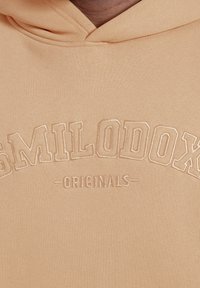 Sudadera beige con el texto bordado "SMILODOX" en letras grandes y "ORIGINALS" debajo en letras más pequeñas en el pecho.