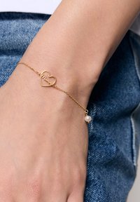 Złota bransoletka z zawieszką w kształcie serca, na której wygrawerowano "Mama". Posiada delikatny łańcuszek oraz mały, okrągły akcent w postaci perły. Noszona na nadgarstku.