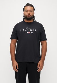 Μαύρο βαμβακερό T-shirt με λευκό λογότυπο "Tommy HILFIGER 1985" και έγχρωμο σημείο με σημαία. Λαιμόκοψη τύπου crew και κοντά μανίκια.