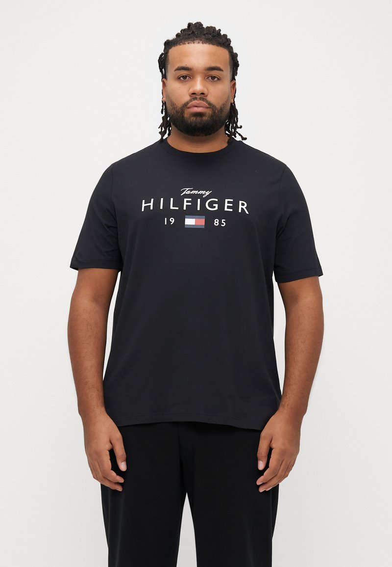 Μαύρο βαμβακερό T-shirt με λευκό λογότυπο "Tommy HILFIGER 1985" και έγχρωμο σημείο με σημαία. Λαιμόκοψη τύπου crew και κοντά μανίκια.