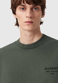 Musta viher puuvillapaita, jossa on ribbi-kaulus ja hillitty logotuloste vasemmassa rinnassa. Pehmeä, tekstuurinen kangas.