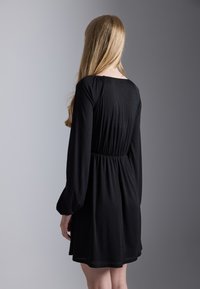 Robe noire à manches longues avec une taille froncée et un tissu doux. Le dos présente un col arrondi et une texture subtile.