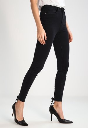 Femme portant un jean skinny noir taille haute avec des détails à lacets aux chevilles et des escarpins noirs à bouts pointus, debout sur un sol blanc.