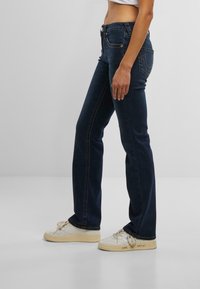 Un model îmbrăcat cu blugi negri și adidași albi stă pe un fundal simplu, evidențiind croiala și designul jeanșilor.