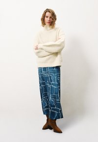 Romig, fluffy trui met hoge hals; teal, gemusterde wijde broek; bruine enkellaarsjes. Model staat met gekruiste armen voor een effen achtergrond.