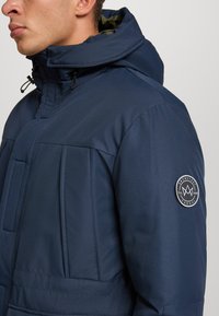 Uomo che indossa un giubbotto imbottito blu scuro con cappuccio e una toppa rotonda bianca e nera sul braccio sinistro.