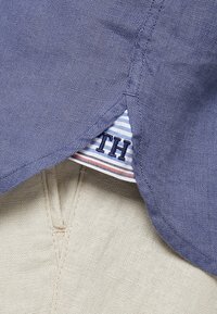 Collar de camisa de lino azul con costura decorativa, superpuesto sobre una tela beige clara, con una etiqueta a rayas con letras en azul oscuro.