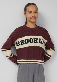 Bordeaux gebreide trui met een crèmekleurige band en de tekst "BROOKLYN", voorzien van gestreepte accenten op de mouwen en een ronde halslijn. Gecombineerd met grijze broek.