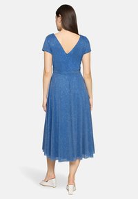 Femme en robe midi bleue scintillante avec un dos en V profond, manches courtes, et sandales blanches à talons, debout devant un fond uni.