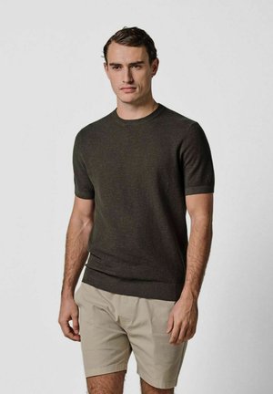 Donkergroene gebreide trui met korte mouwen en een ronde hals, gecombineerd met lichtbeige korte broek. Glad oppervlak, minimalistisch ontwerp.