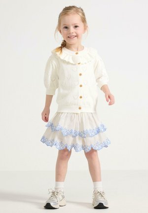 Weißer Strickcardigan mit Herzmustern und Rüschkragen, kombiniert mit einem geschichteten cremefarbenen Rock, der mit blauen Blumenstickereien verziert ist.