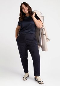 Marinblus i satin med rund halsringning, kombinerad med mörka byxor med framfickor, och sneakers i krämvit och marinblå. Håller en ljus trenchcoat.