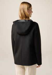 Abrigo negro con capucha y una silueta estructurada. Hecho de tejido texturizado, con un forro interior suave y un diseño limpio en la parte posterior.