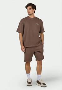 Ruskea ribbineulottu t-paita lyhyillä hihoilla, jossa on pieni logo, yhdistettynä samanlaisiin shortseihin. Yhdistetty valkoisiin ja harmaisiin tennareihin ja sukkiin.