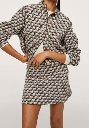 Femme portant une veste à motifs et une jupe assortie avec un motif géométrique en forme de cube dans des nuances de gris, bleu et marron sur un fond uni.