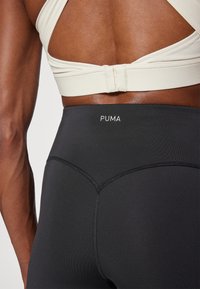 Juodos leggingai su aukštu liemeniu ir dekoratyviniais siūlais, su baltu "PUMA" logotipu ant nugaros. Slydus, tamprus audinys.