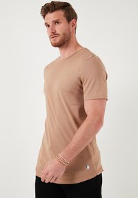 Světlé hnědé tričko s krátkým rukávem vyrobené z hladkého materiálu, s kulatým výstřihem a malou značkovou nášivkou na lemu.