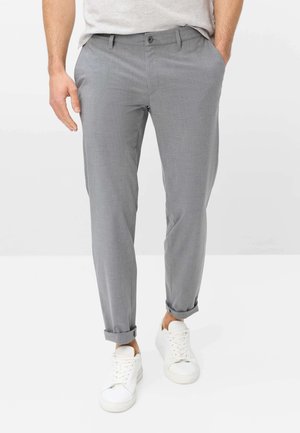 Mand iført lysegrå skinny bukser rullet op ved anklerne og hvide sneakers, med den ene hånd i lommen og neutral baggrund.