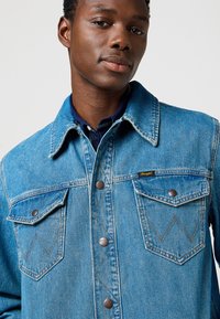 Chaqueta de mezclilla azul claro con un collar puntiagudo, botones de presión de metal y dos bolsillos en el pecho que presentan un patrón de puntada en zigzag.