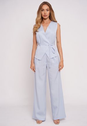 Kobieta o długich blond włosach, ubrana w jasnoniebieski top bez rękawów z kopertowym dekoltem i pasujące szerokie spodnie, stojąca na jednolitym tle.