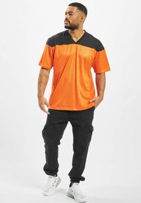 Oranži ja must lühikeste varrukatega spordipari, millel on V-kaelus, koos mustade cargo-pükstega ja valgete tossudega. Sile, hingav kangas.