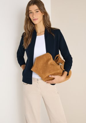 Donna con giacca blu navy, camicia bianca, pantaloni beige chiaro, che tiene una grande borsa in suede marrone su uno sfondo uniforme.