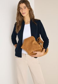 Femme portant une veste bleu marine, une chemise blanche, un pantalon beige clair, tenant un grand sac à main en daim marron sur un fond uni.