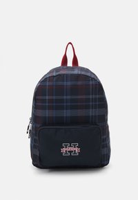 Zaino a quadri in blu navy, rosso e blu con una tasca con zip e una patch con logo. Presenta due spallacci in tessuto rosso nella parte superiore. Materiale testurizzato.