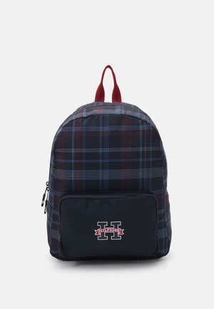 Zaino a quadri in blu navy, rosso e blu con una tasca con zip e una patch con logo. Presenta due spallacci in tessuto rosso nella parte superiore. Materiale testurizzato.