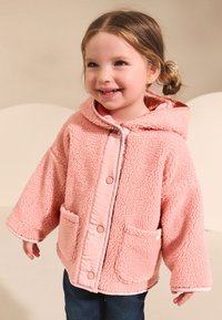 Różowa polarowa kurtka z kapturem, dużymi kieszeniami i zatrzaskami. Miękka faktura, szerokie rękawy oraz kontrastowe obszycie na krawędziach.